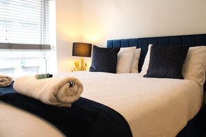 Ein ordentlich gemachtes Bett mit blauem Kopfteil, einer Nachttischlampe und einem Fenster mit Jalousien.
