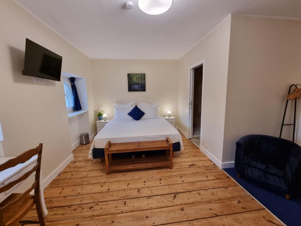 Quarto com cama, mesas de cabeceira, televisão, pintura, um banco de madeira e um sofá azul.