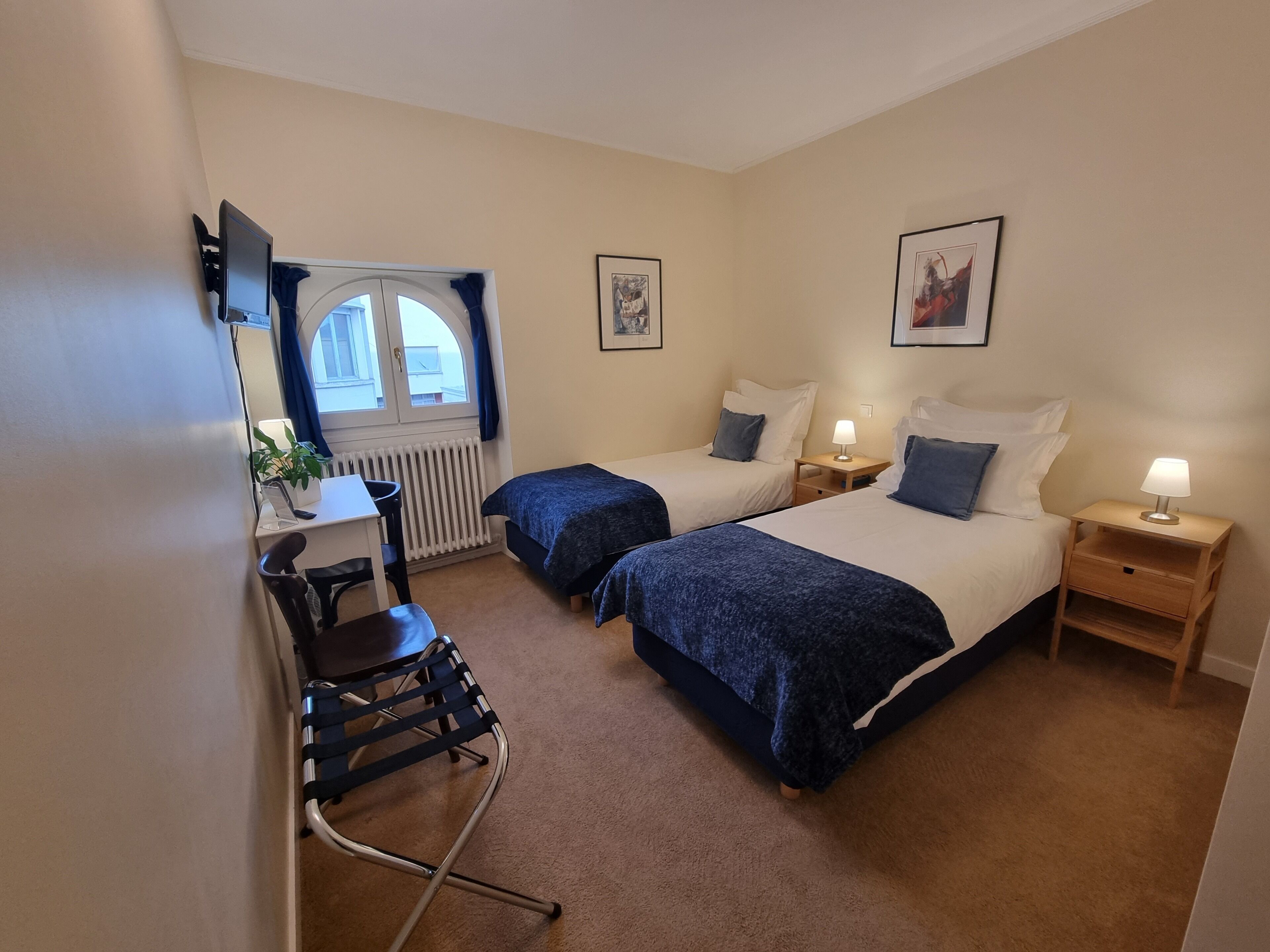A hotel room with two beds, a desk, a chair, a radiator, and a window with curtains.