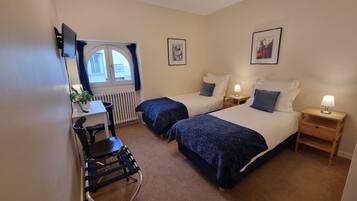 A hotel room with two beds, a desk, a chair, a radiator, and a window with curtains.