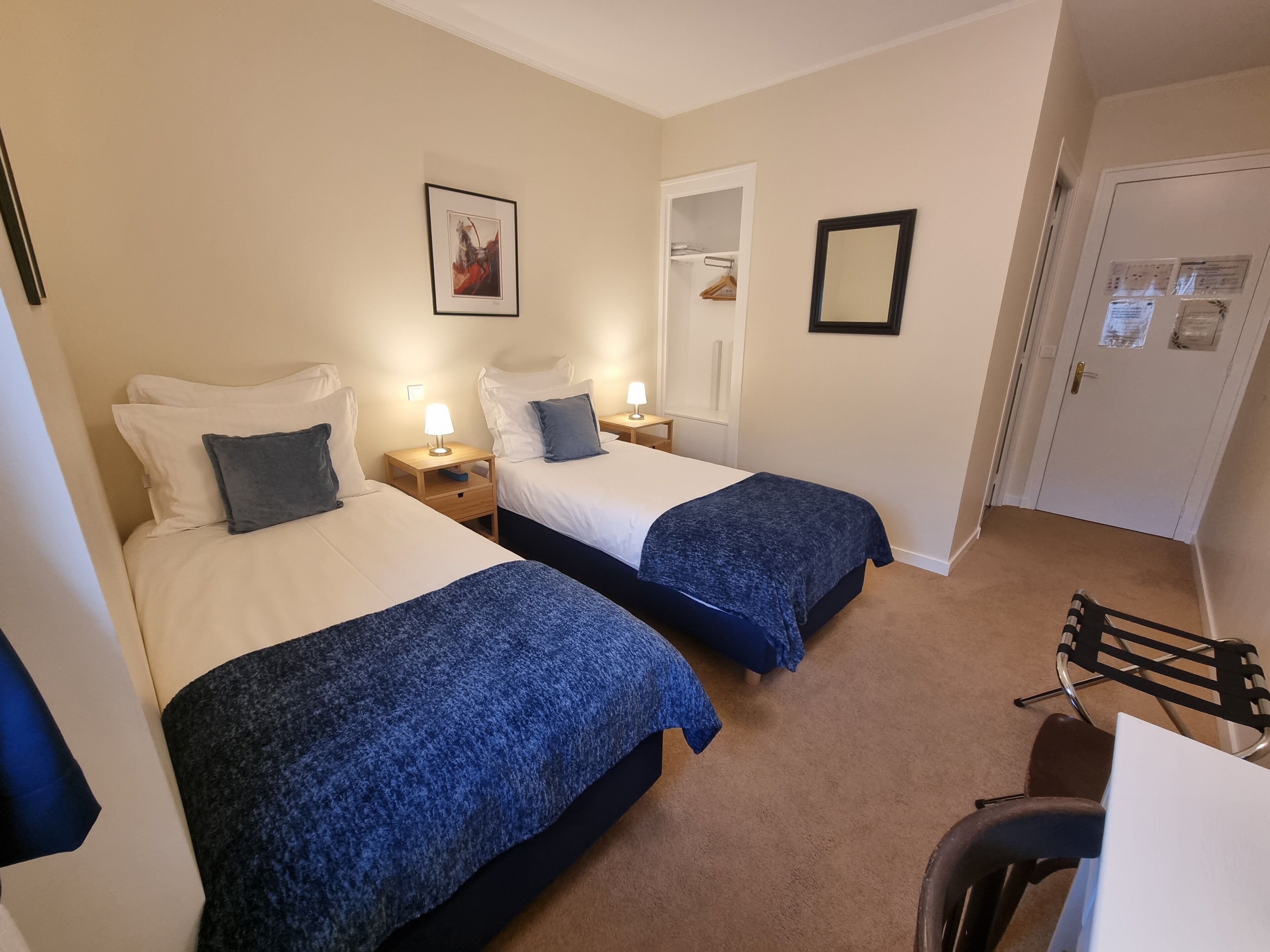 A hotel room with two beds, a desk, a chair, a mirror, and a door with a bulletin board.