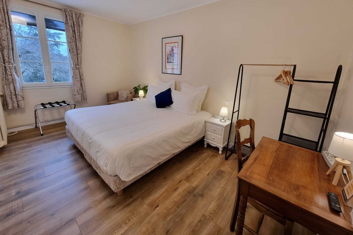 A bedroom with a bed, bedside table, lamp, chair, and a wooden table with a remote control.