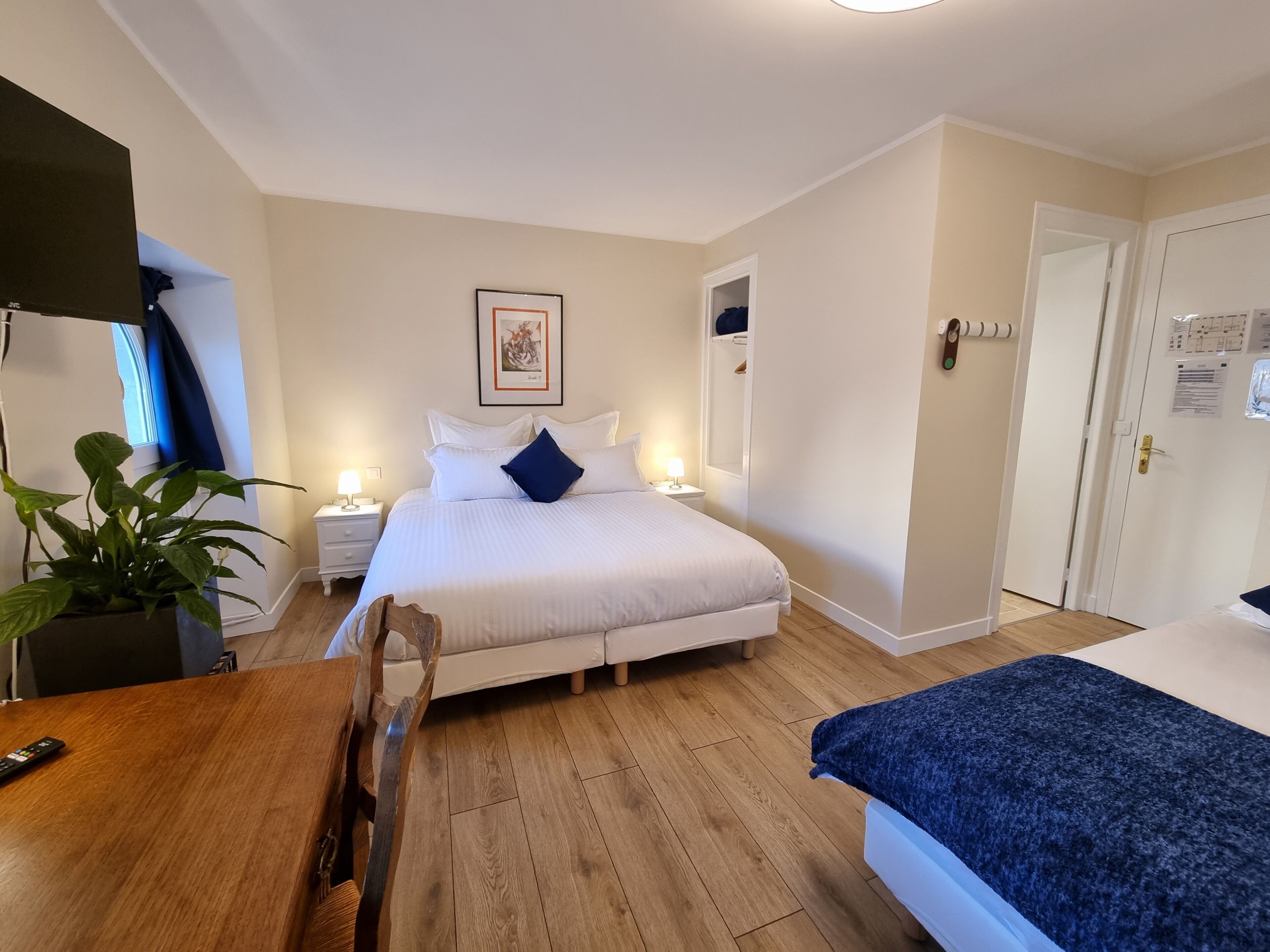 Ett hotellrum med en säng, sängbord, ett skrivbord, en stol, en krukväxt och en dörr.
