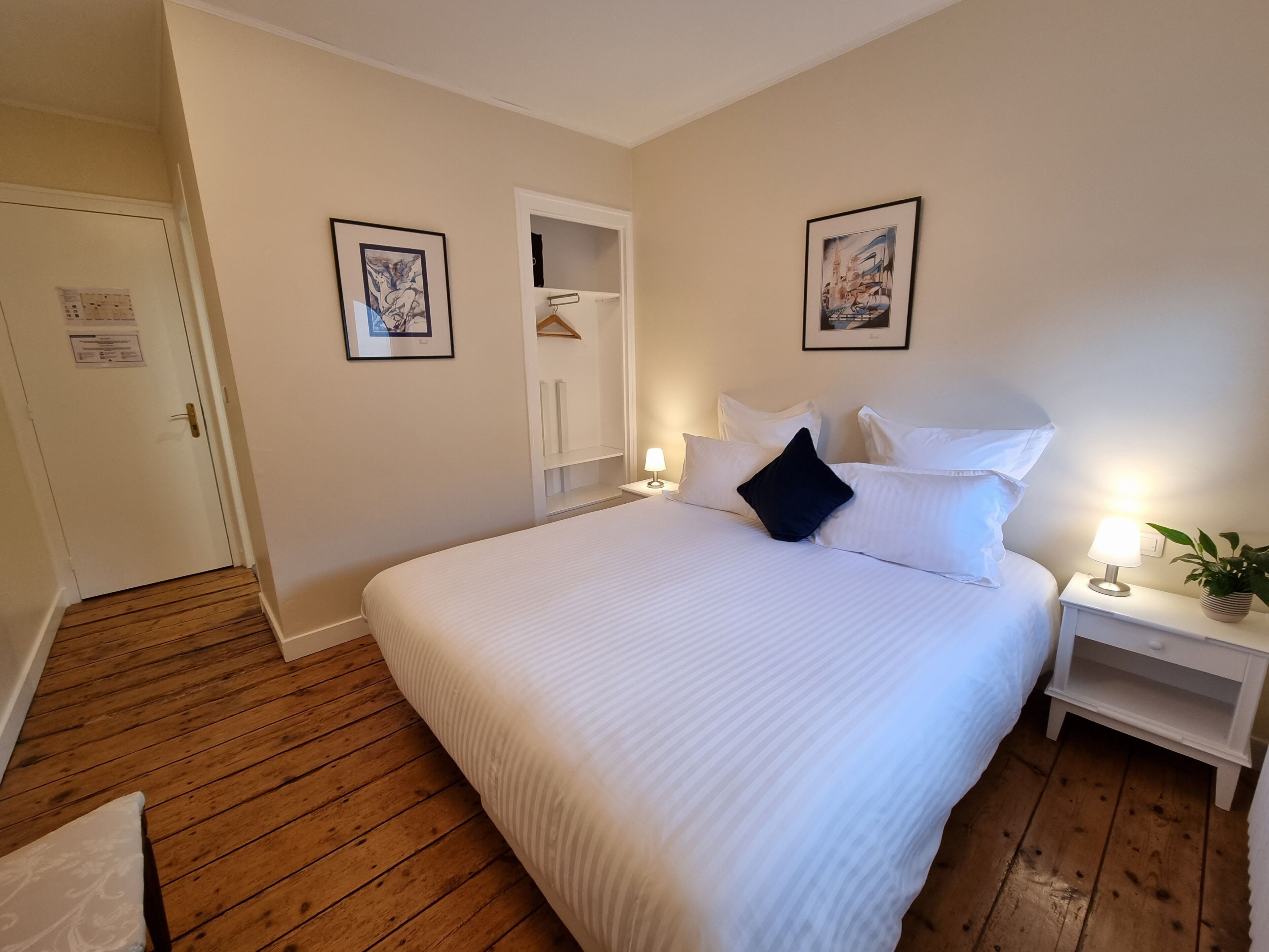 A hotel room with a large bed, bedside tables, a wardrobe, and framed artwork on the walls.