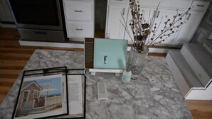En køkkenbordplade med et rammet fotografi, en bog med titlen 'A guest book', en vase med visne blomster og en lille elektronisk enhed.