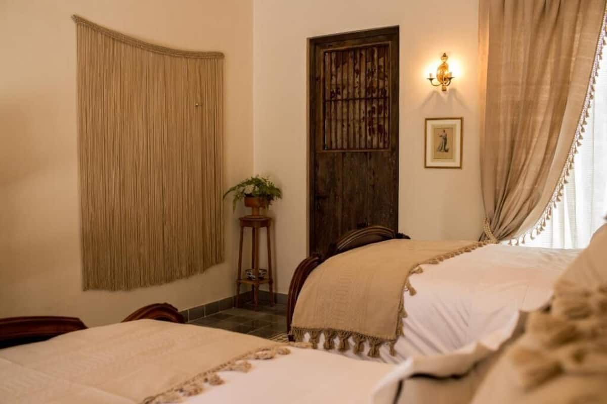 A hotel room with two beds, a wooden door, a wall-mounted light fixture, and a window with curtains.