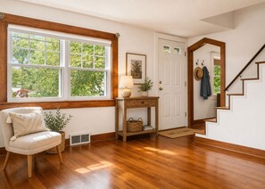 A room with hardwood flooring, a chair, a table with a lamp, a door, and a staircase. - Charming 1940s Cape Cod Retreat on 1 Acre - Fireplace & Large Yard (Ashland)