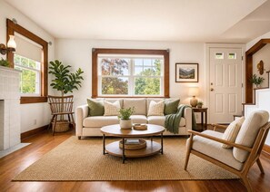 A living room with a sofa, coffee table, and chairs, a fireplace, a large window with a view, and a door. - Charming 1940s Cape Cod Retreat on 1 Acre - Fireplace & Large Yard (Ashland)