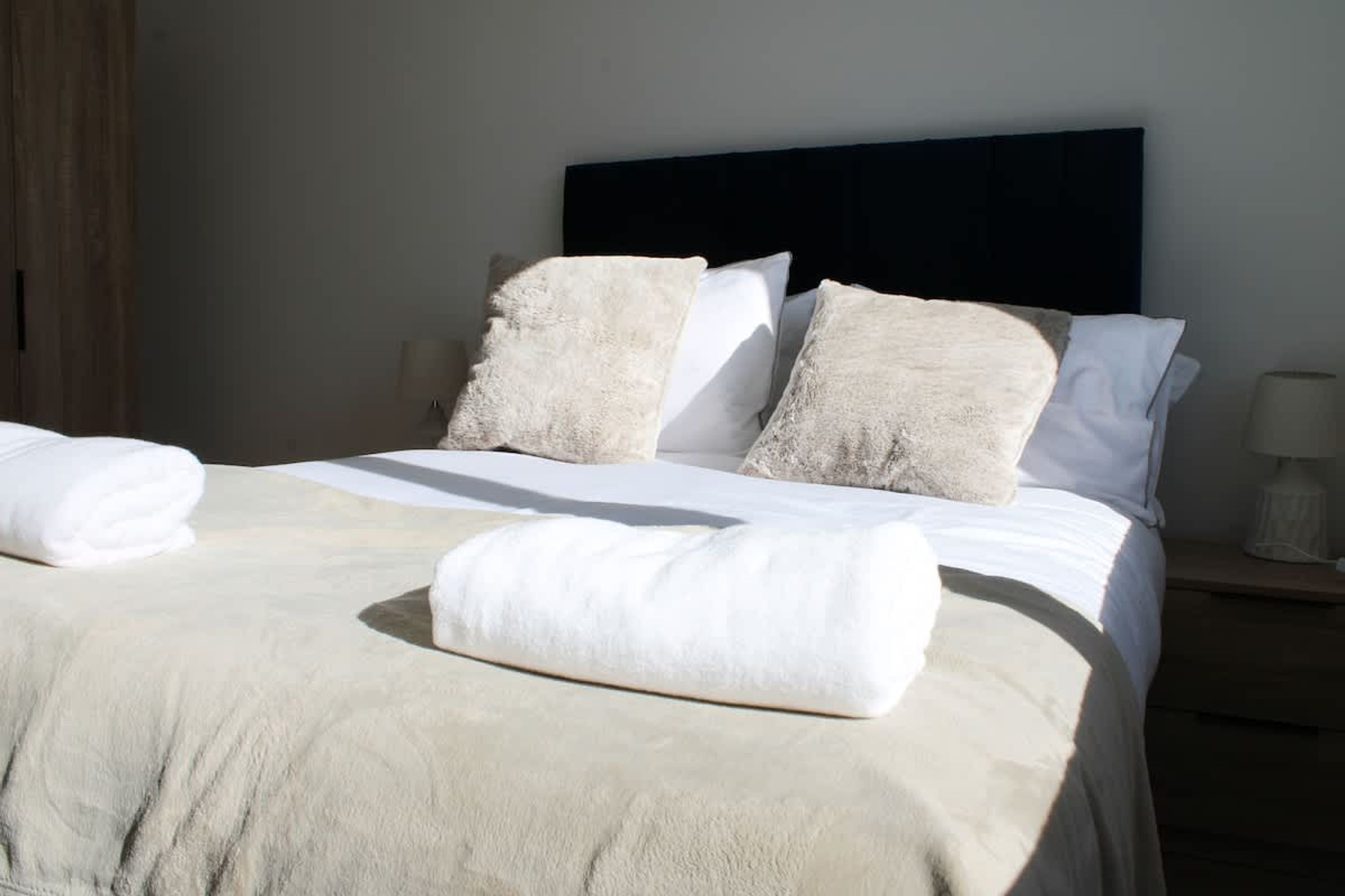 Un lit bien fait, avec une grande couette beige et des oreillers, une table de chevet en bois avec une lampe, et une tête de lit sombre.