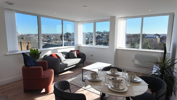 A modern living room with a sofa, chairs, a dining table set for four, and large windows offering a city view. - NEW Modern Apartment Free Parking Workspace (Gravesend)
