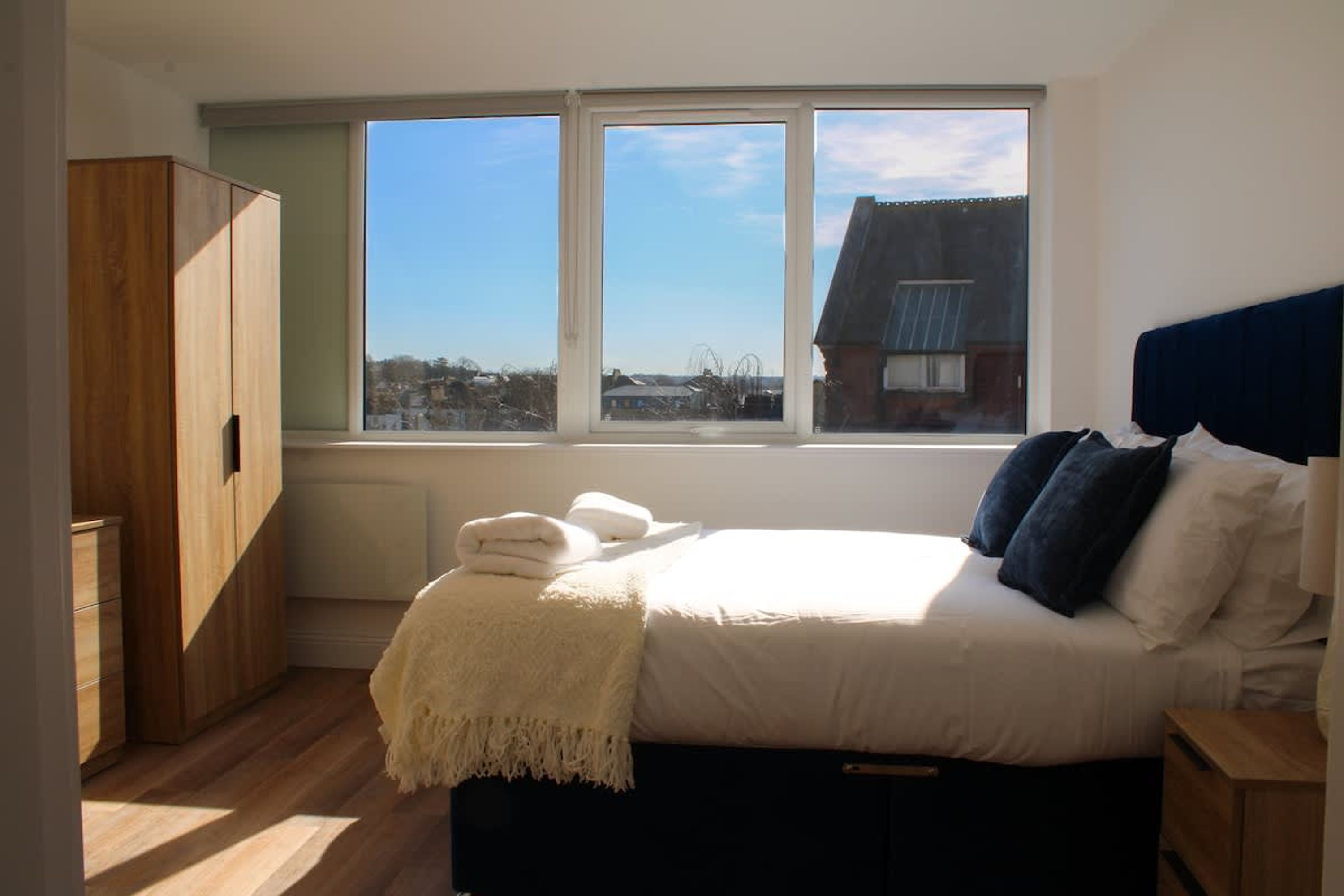 Een slaapkamer met een groot raam, een bed met kussens, een nachtkastje en een kledingkast.