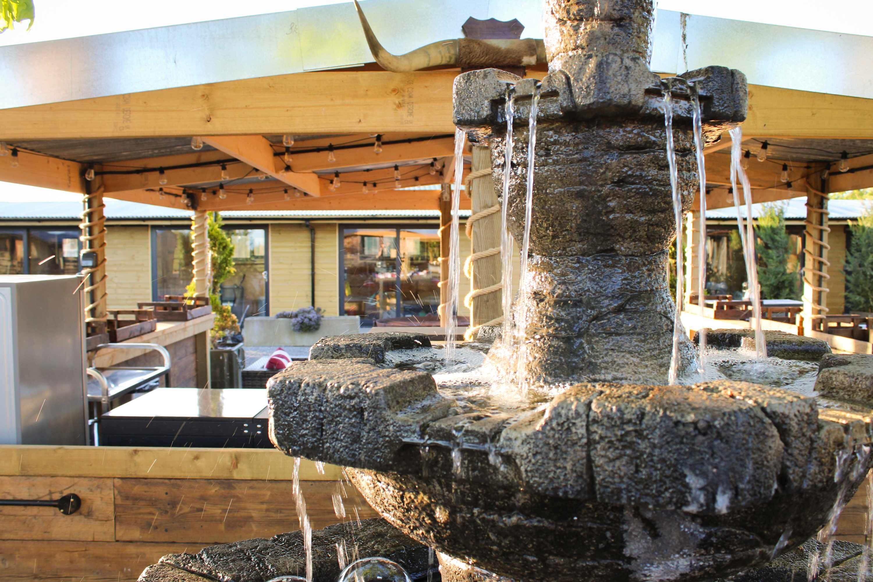 A stone fountain with water flowing from multiple spouts, a wooden pergola overhead, and a grill area in the background.