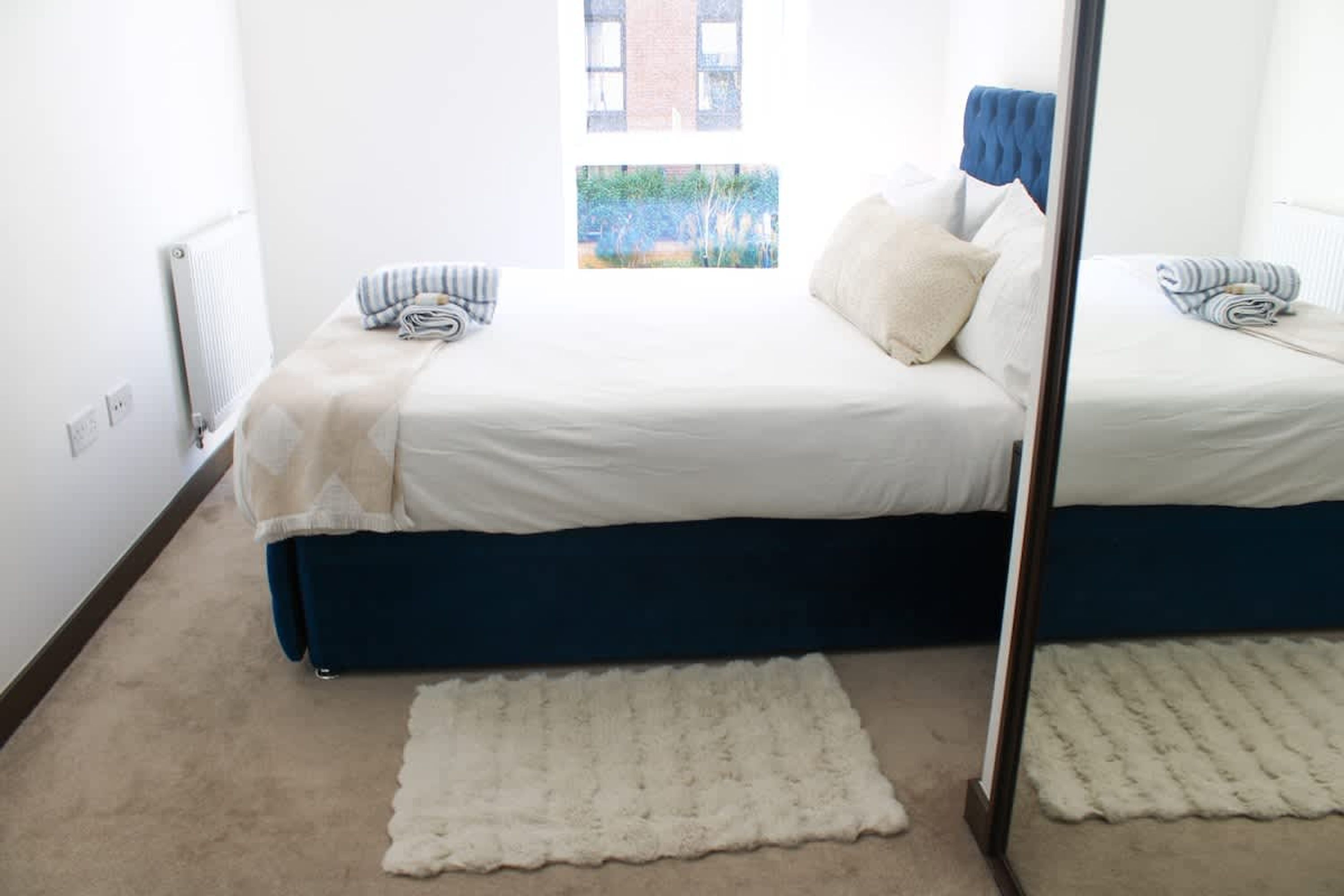 Een slaapkamer met een tweepersoonsbed, een raam met uitzicht op een gebouw, en een wit vloerkleed.