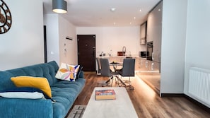 A modern living room with a blue sofa, a dining table with chairs, a kitchen area with a microwave, and a door leading to another room. - The Ivy Abode Stylish 2-bed Apartment Parking (Harlow)