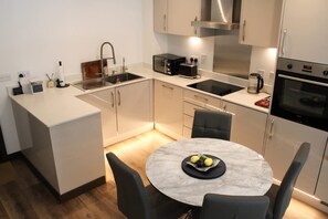 A modern kitchen with a built-in stove, microwave, and sink, a round dining table with chairs, and a small island with a sink. - The Ivy Abode Stylish 2-bed Apartment Parking (Harlow)