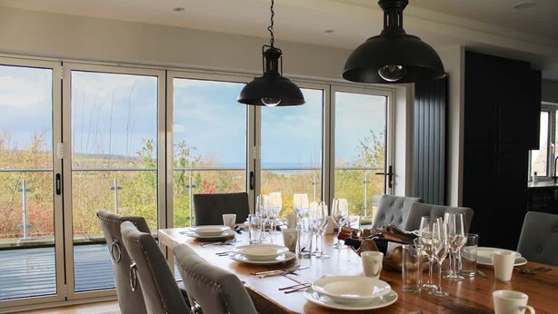 A dining area with a large table set for a meal, large windows with a view, and modern pendant lighting. - Luxury Sea View Home With Hot Tub Sauna (Llanerchymedd)