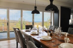 A dining area with a large table set for a meal, large windows with a view, and modern pendant lighting. - Luxury Sea View Home With Hot Tub Sauna (Llanerchymedd)
