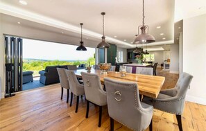 Een eetgedeelte met een houten tafel en grijs gestoffeerde stoelen, een keuken op de achtergrond en uitzicht op groen door grote ramen.