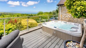 A wooden deck with a hot tub, a stone wall, and a view of the landscape. - Luxury Sea View Home With Hot Tub Sauna (Llanerchymedd)