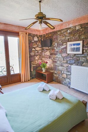 Un dormitorio con cama, ventilador de techo, pared de piedra, televisor, radiador y ventana con cortinas.