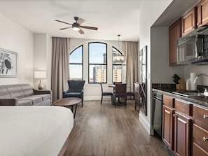 A hotel room with a bed, a seating area with a table and chairs, a kitchenette, and large windows with a city view.