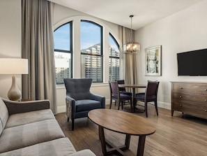 A hotel room with a sofa, a dining table with chairs, a TV, and large windows with curtains.