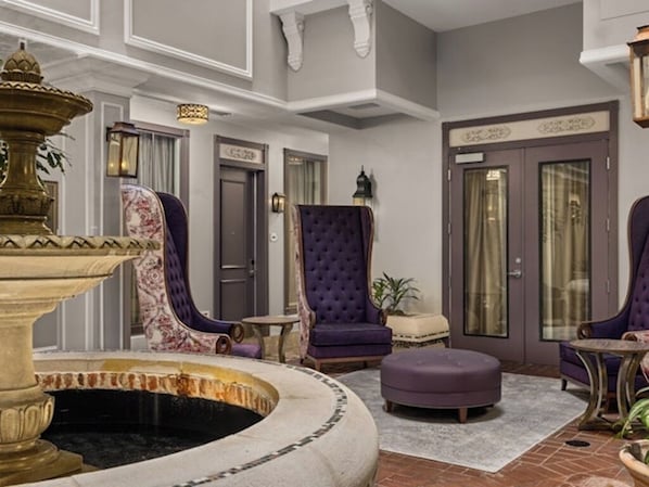 A lobby with a fountain, purple upholstered chairs, a round ottoman, and a door with a sign.