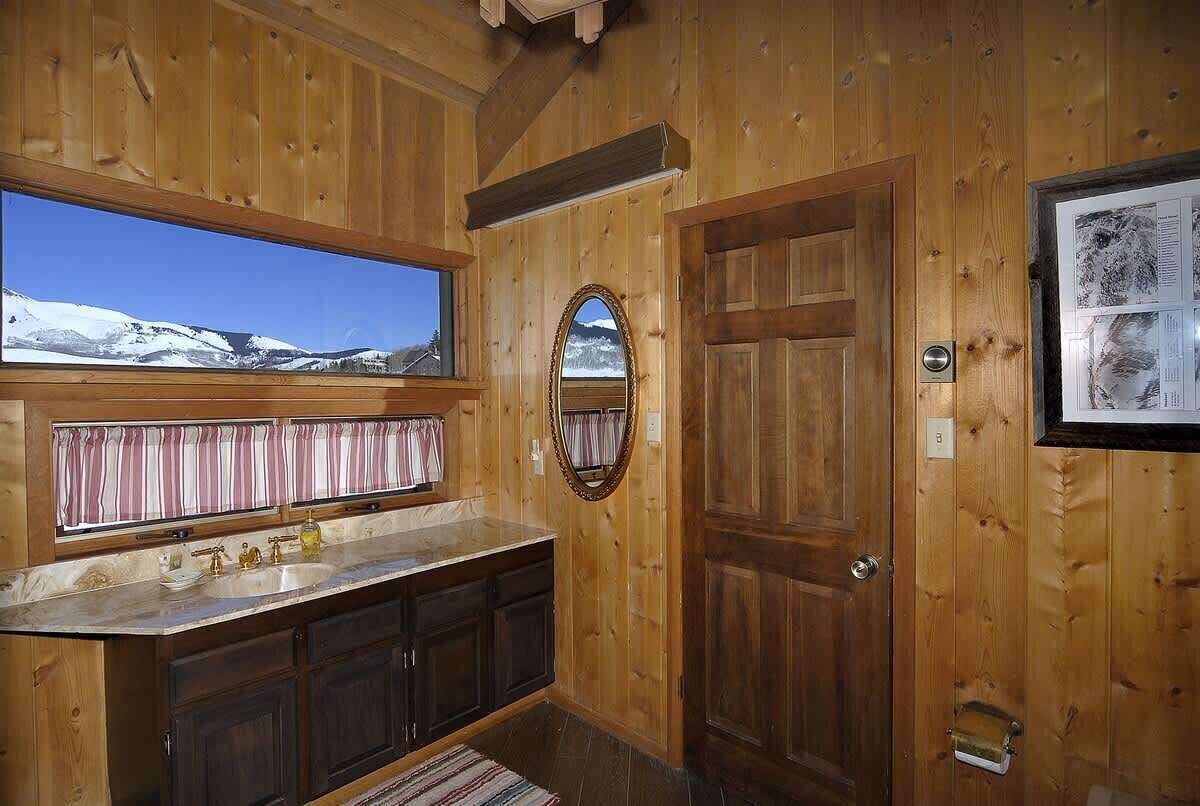 A wooden cabin bathroom with a large window, a sink, cabinets, and a door.