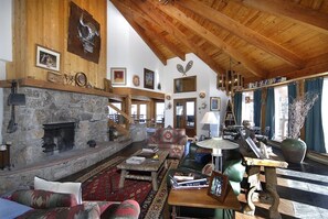 Ein rustikales Interieur mit Kamin aus Stein, Holzbalken und verschiedenen Sitzbereichen, darunter eine Couch und ein runder Tisch.