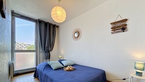 Ett sovrum med en säng, ett fönster med gardiner, en spegel som hänger på väggen, ett litet bord med en lampa och en väggdekoration med text.