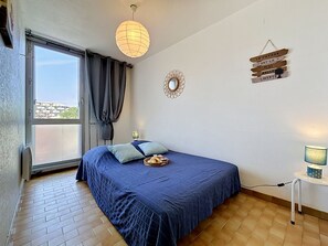 Ein Schlafzimmer mit Bett, einem Fenster mit Vorhängen, einem Wandspiegel, einem kleinen Tisch mit Lampe und einer Wanddekoration mit Text.