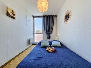 A bedroom with a bed, a window with curtains, a wall-mounted lamp, and a decorative mirror. - La Rose des Sables by Interhome (La Grande Motte)