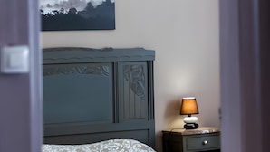 A bedroom with a bed, a nightstand with a lamp, a wall-mounted painting, and a door frame.