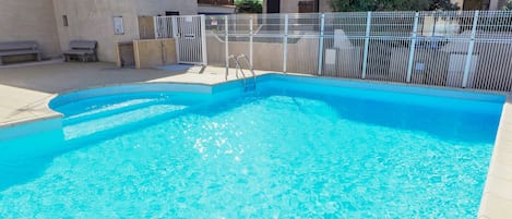A clear blue swimming pool with a metal fence, benches, and a building in the background.
