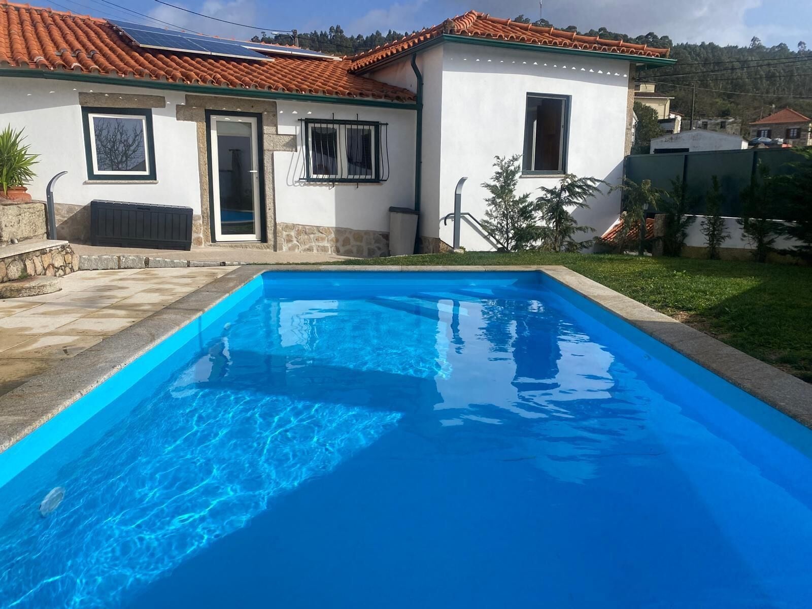 Une piscine avec une terrasse carrelée, une maison avec un toit recouvert de tuiles rouges et un mur en pierre.