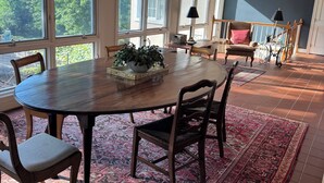 Uma área de refeições com mesa redonda, cadeiras, um candelabro e vista para árvores através de grandes janelas.