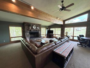 A spacious living room with a stone fireplace, leather sofa, and large windows offering a view of the outdoors. - King Davis House-Best Views in Mt. Crested Butte (Crested Butte)