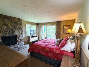 A bedroom with a stone wall, a bed with a red bedspread, a wooden dresser, a lamp, and a window with curtains. - King Davis House-Best Views in Mt. Crested Butte (Crested Butte)