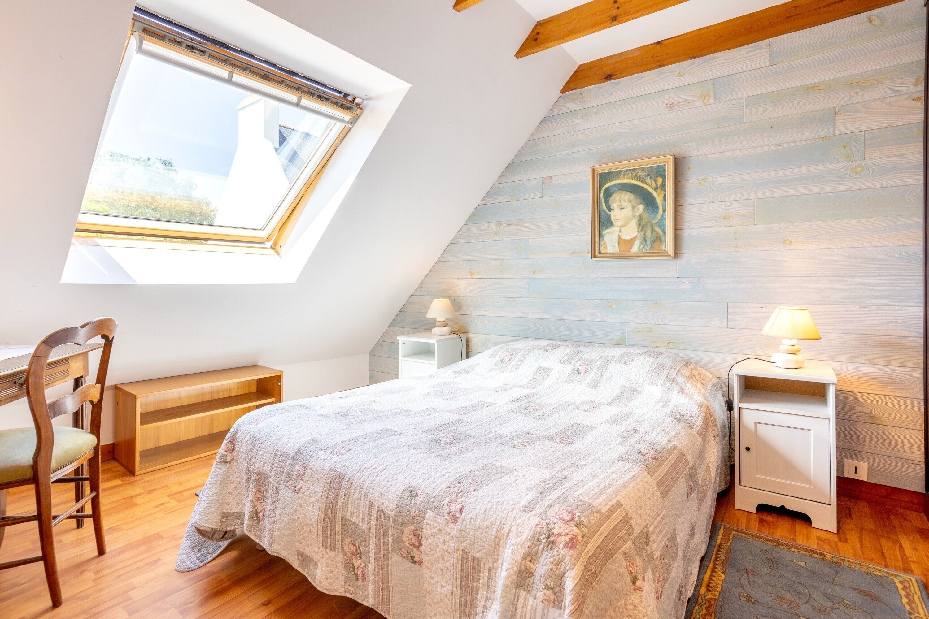 A bedroom with a sloped ceiling, a bed with a floral bedspread, a nightstand with a lamp, a wooden chair, and a picture on the wall.