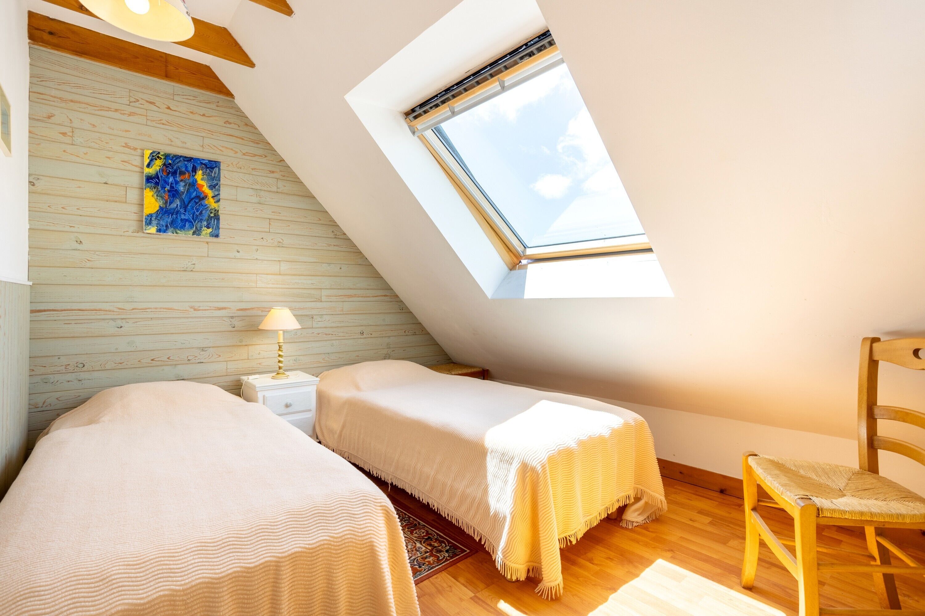 A room with a sloped ceiling, two beds, a chair, a nightstand, a lamp, a painting, and a skylight.