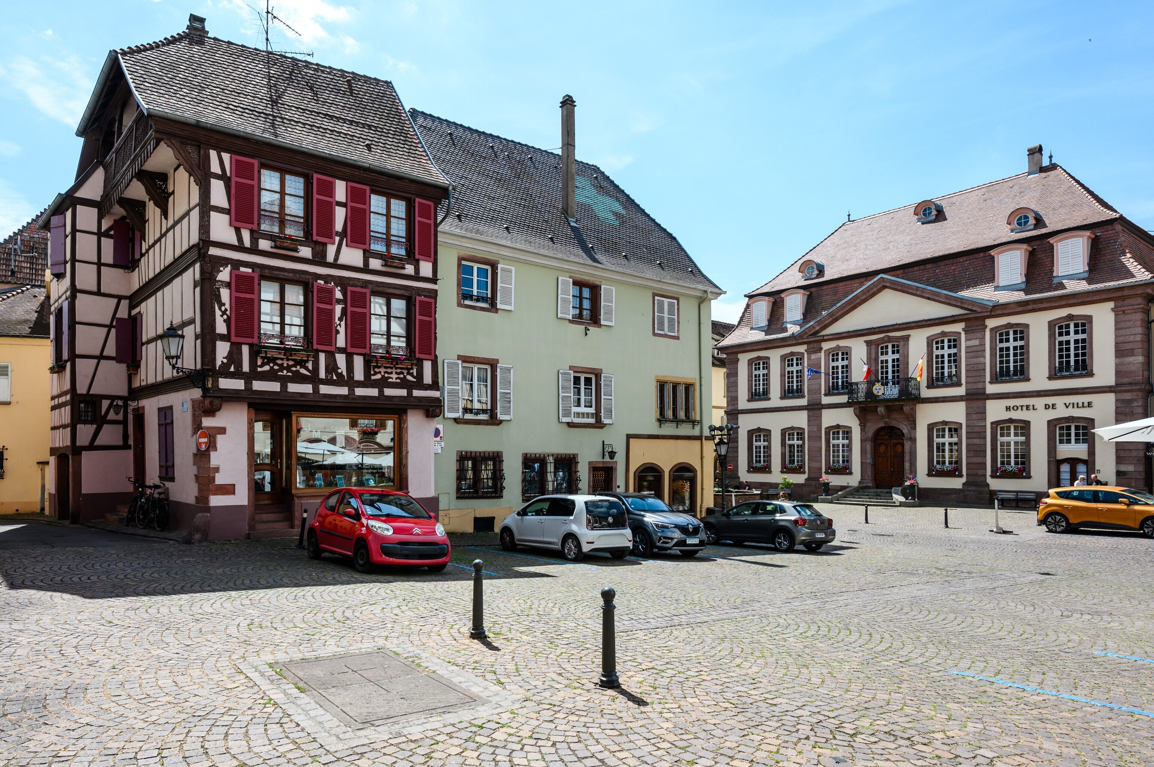 Une place pavée bordée de bâtiments historiques, une voiture rouge et une voiture blanche garées devant un bâtiment portant l’inscription « HÔTEL DE VILLE ».