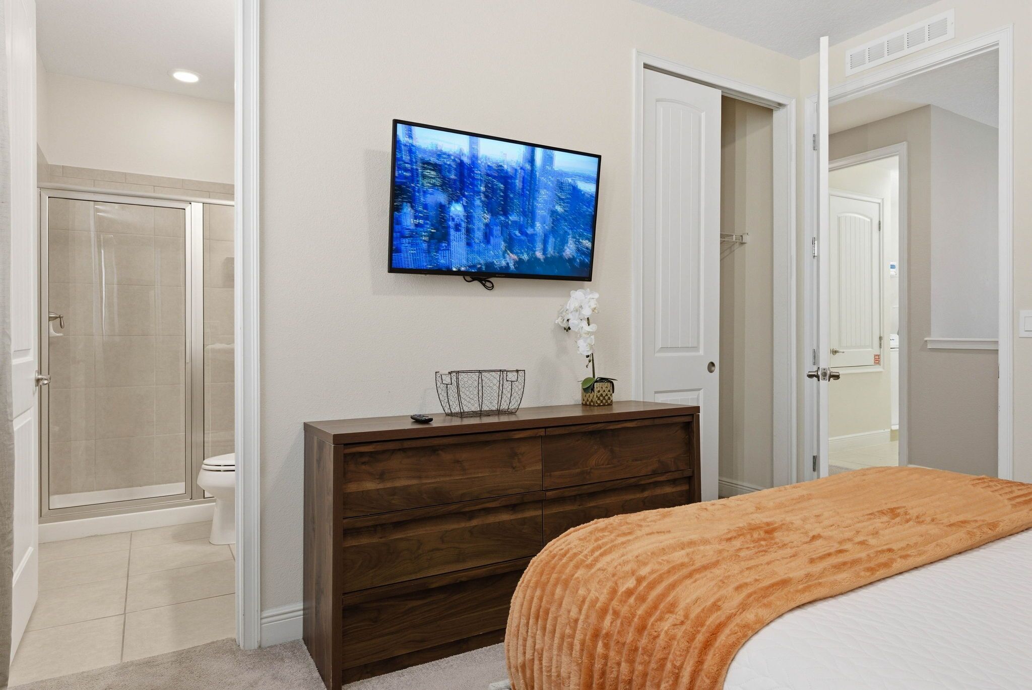 A bedroom with a bed, a TV mounted on the wall, a wooden dresser, and a shower area.