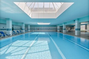 Une piscine intérieure à l’eau d’un bleu cristallin, avec un plafond baigné de lumière et des chaises longues bleues disposées le long du bord.