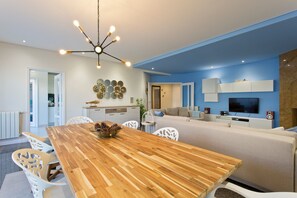 A modern living room with a wooden dining table, white chairs, a sofa, a TV, and a ceiling light fixture. - Beach Sunset – Oceanfront Apartment with Terrace and Sea View (Vila Nova de Gaia)