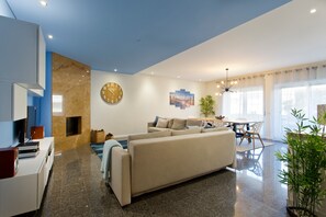 A modern living room with a sofa, a coffee table, a dining area with chairs, a clock on the wall, and a plant. - Beach Sunset – Oceanfront Apartment with Terrace and Sea View (Vila Nova de Gaia)
