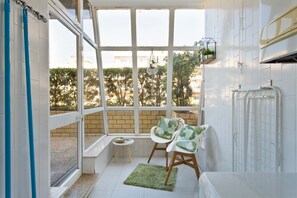A room with a large window, a small table, two chairs, a radiator, and a green rug. - Beach Sunset – Oceanfront Apartment with Terrace and Sea View (Vila Nova de Gaia)