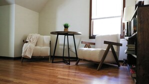 A room with a wooden floor, a white armchair, a round table with a plant, and a bookshelf. - Country Oasis 4-bedroom farmhouse in tranquil Shelby with cozy charm (Shelby)