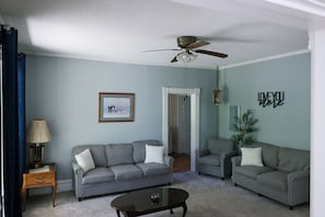 A living room with a grey sofa, a coffee table, a ceiling fan, and a wall with a 'LOVE YOU MORE' sign.