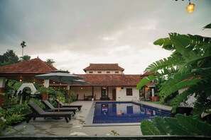 A pool area with lounge chairs, umbrellas, and a building with a tiled roof.
