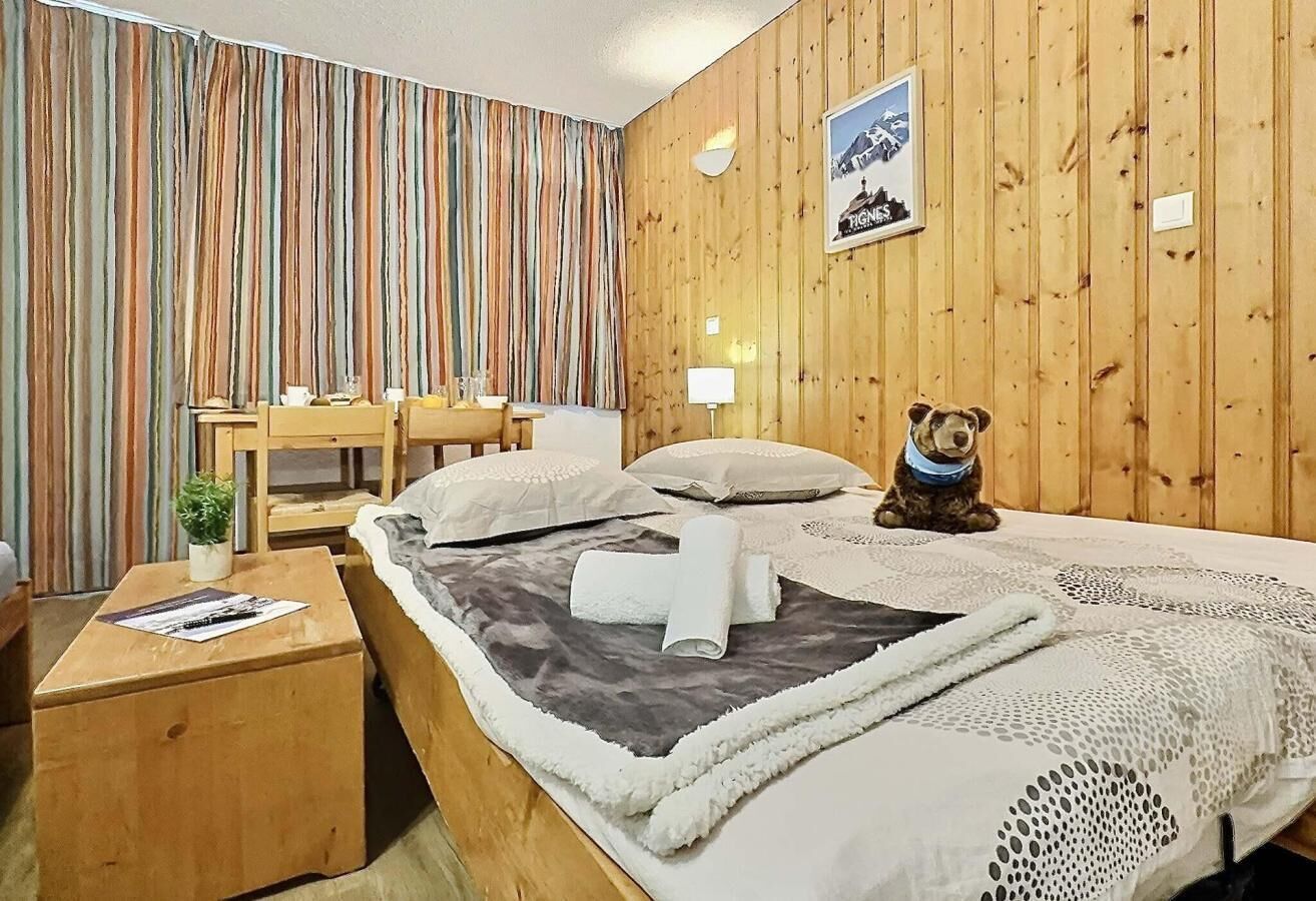 Et hotelværelse med væg i træpaneler, en seng med gråt sengetæppe, et lille bord med en plante og en bamsefigur på sengen.